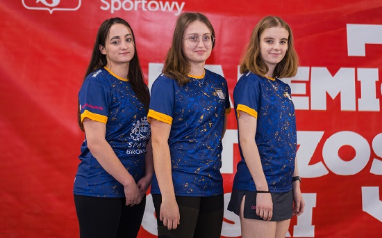 trzy dziewczyny w strojach sportowych pozują do zdjęcia na tle ścianki z nazwą mistrzostw