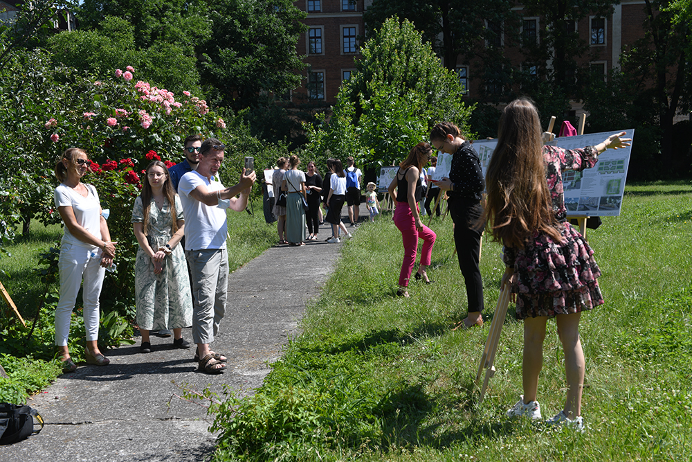 Wzdłuż ścieżki ustawione są plansze z pracami studentów. Duża grupa ludzi spaceruje i ogląda wystawę.