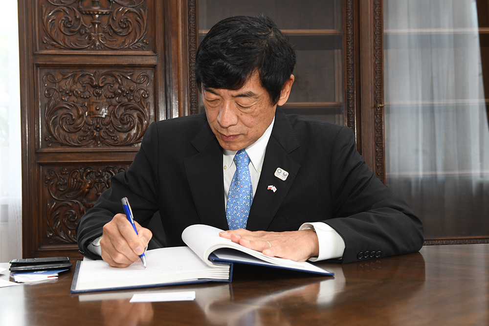 Ambasador Japonii siedzi przy stole. Pochyla się nad książką, do której coś wpisuje. W tle znajdują się eleganckie meble z ciemnego drewna. 