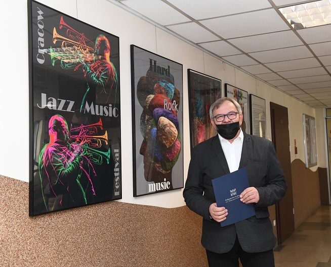 Profesor Zbigniew Latała stoi obok ściany na której wiszą jego prace składające się na wystawę „Nie tylko JAZZ, not only”