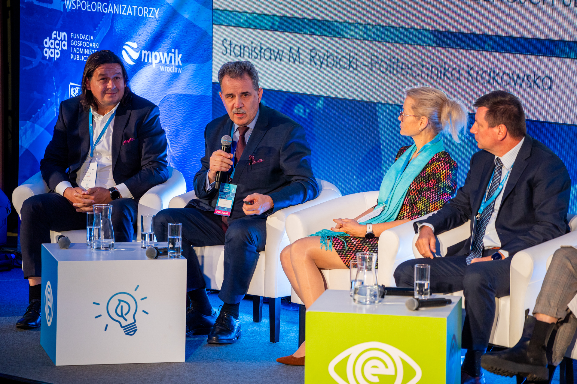 Cztery osoby siedzą na scenie i prowadzą dyskusję. Wśród nich profesor Stanisław Rybicki, który trzyma mikrofon. 