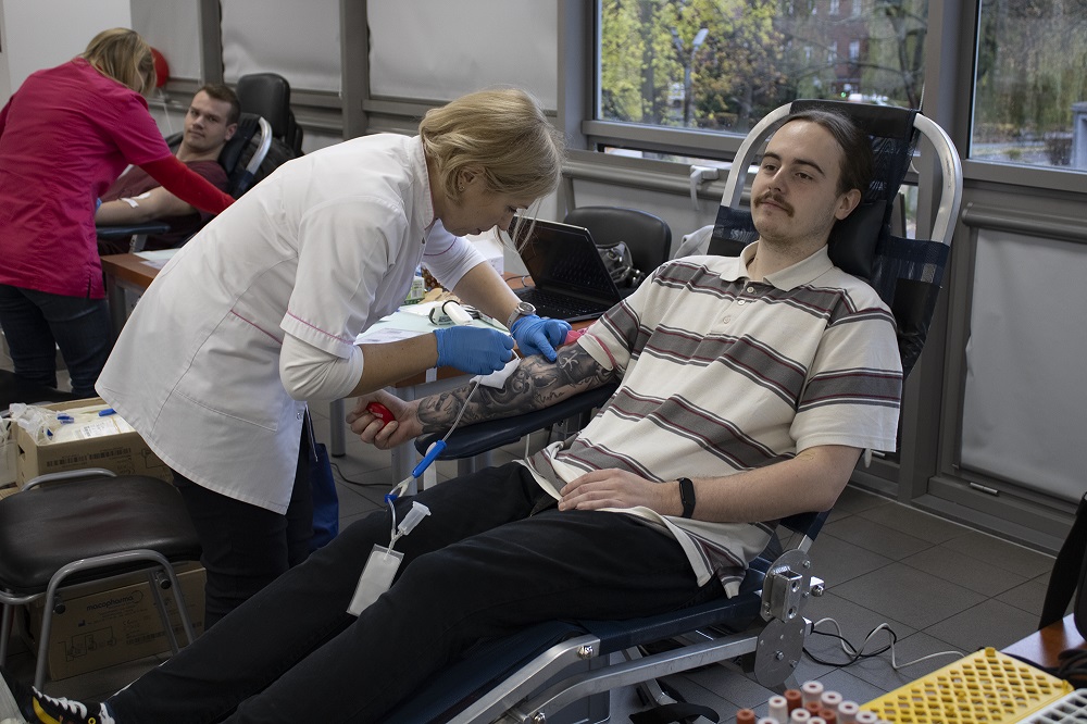 Pielęgniarka w białym kitlu pochyla się nad ręką siedzącego na fotelu do pobierania krwi mężczyzny