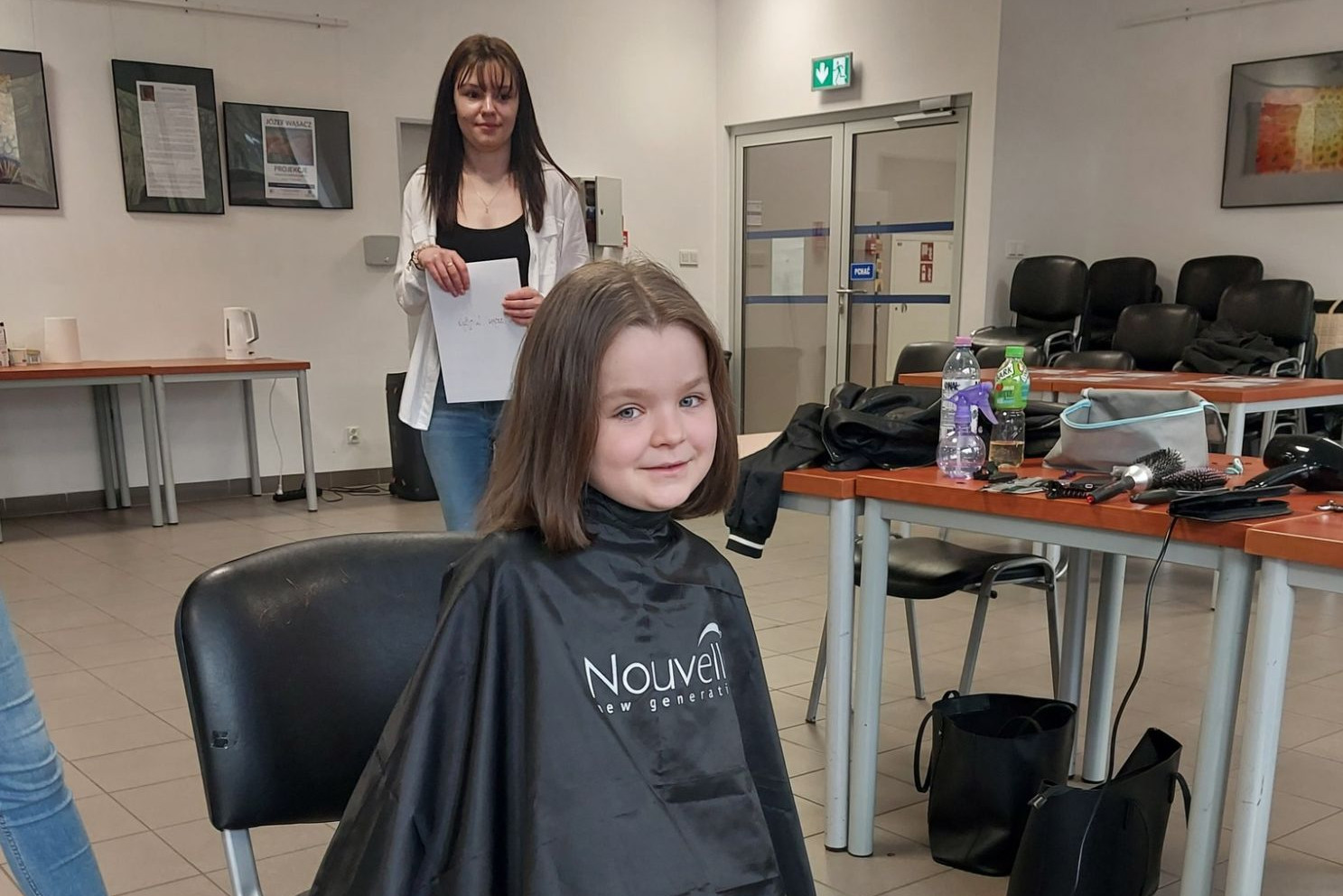 Dziewczynka z krótkimi włosami ubrana w pelerynę fryzjerską. Prezentuje ona nową fryzurę po oddaniu włosów w ramach akcji "o włos od pomocy"