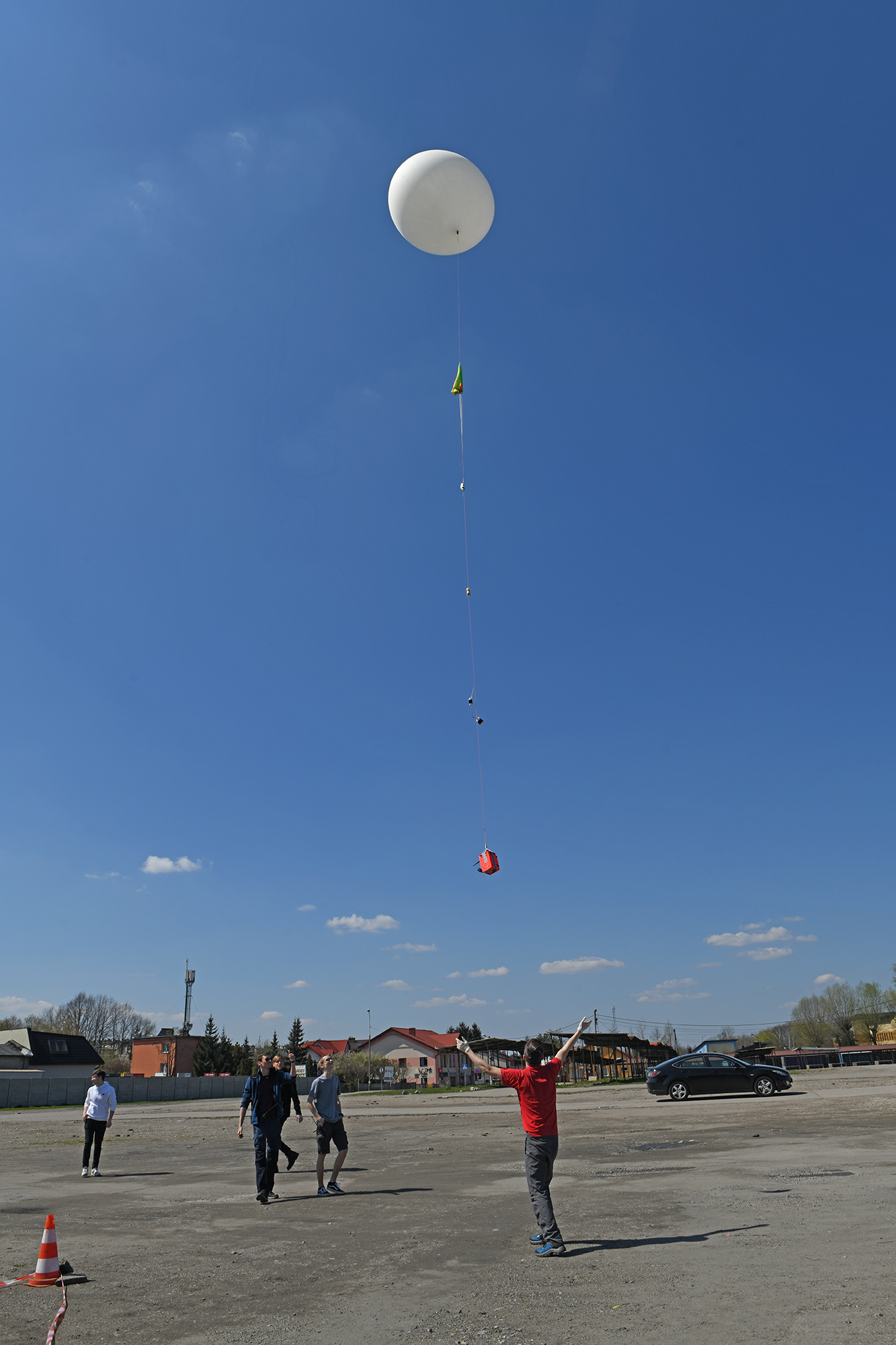 Balon z sondą unosi się w powietrze. Na ziemi stoją członkowie koła naukowego i spoglądają do góry, przyglądając się balonowi.