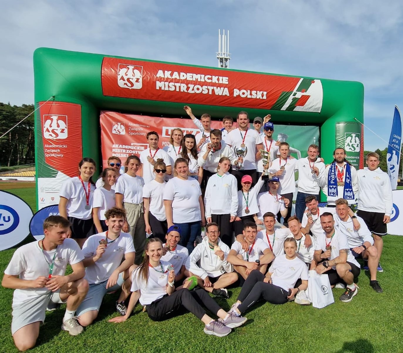 Grupa radosnych ludzi w białych bluzach i t-shirtach. Za nimi napis Akademickie Mistrzostwa Polski.