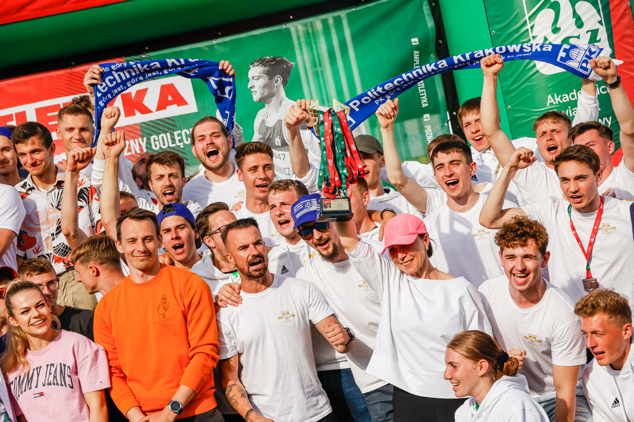 Grupa radosnych młodych ludzi wznosi w górę medale i szaliki z napisem "Politechnika Krakowska, górą jest"