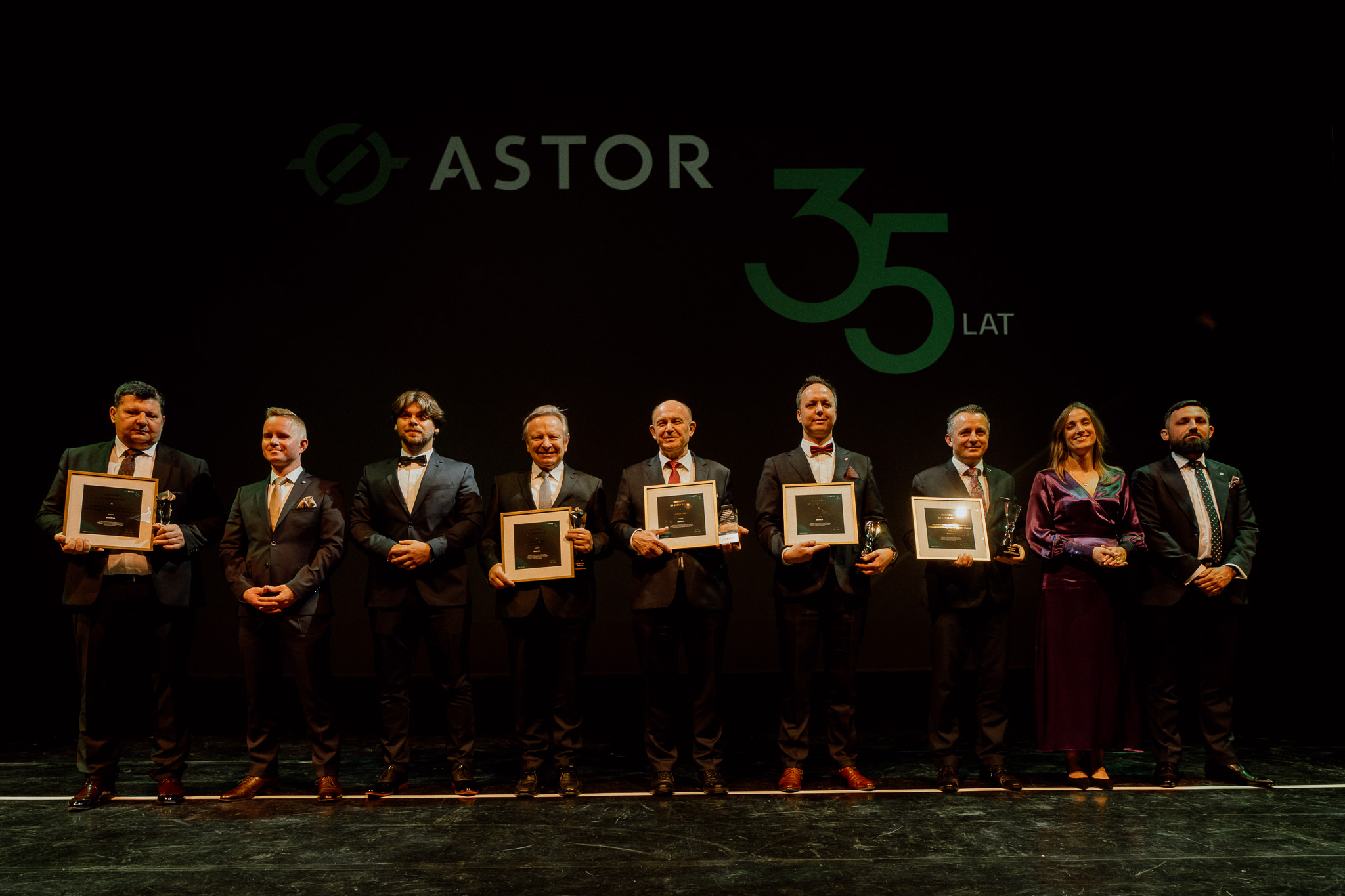 Ośmiu mężczyzn w garniturach i jedna blondynka w długiej fioletowej sukni. Stoją na scenie. Wśród nich jest prof. Sładek. Część osób trzyma w rękach dyplomy. W tle logotyp firmy ASTOR i napis „35 lat”. 