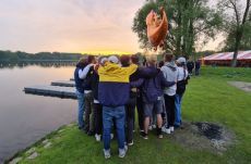 Grupa młodych ludzi stoi nad brzegiem jeziora. Zwróceni są tyłem do obiektywu. Stoją w okręgu i obejmują się. Nad nimi unosi się pomarańczowy balonik w kształcie smoka. W jeziorze odbija się zachodzące słońce.