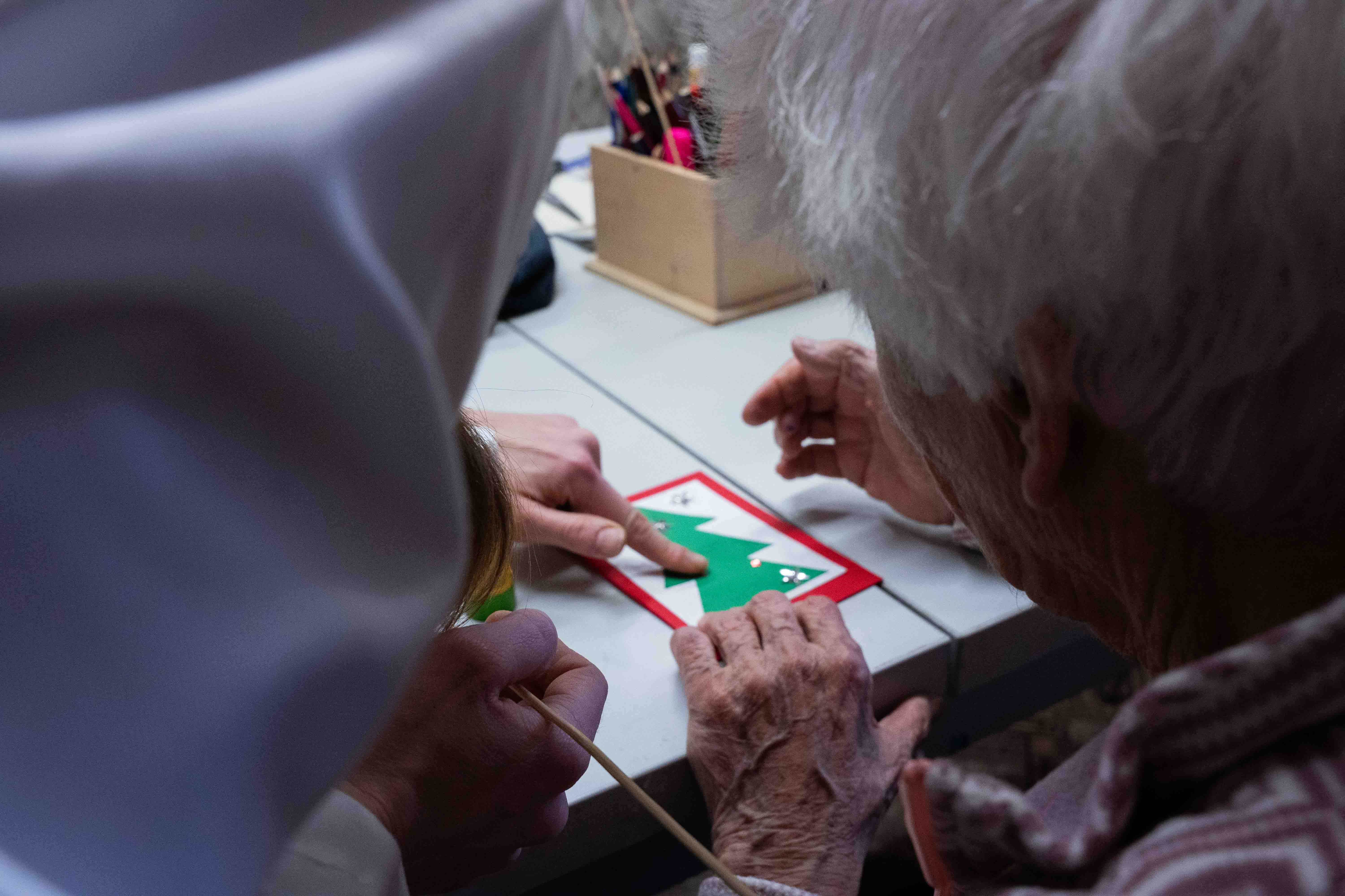 Dłonie starszej i młodej osoby pracują wspólnie nad kartką świąteczną, na której widoczna jest choinka wycięta z papieru. 