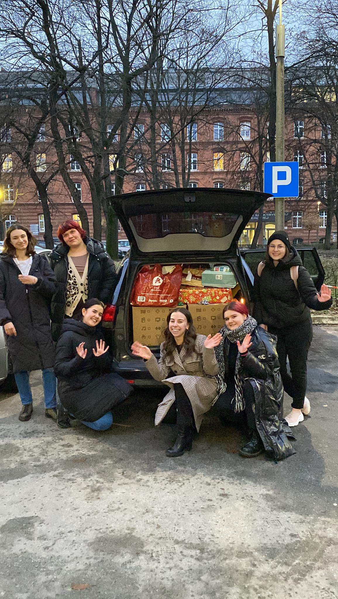 Grupa młodych ludzi pozuje do zdjęcia przy otwartym bagażniku samochodowym, który wypełniony jest prezentami