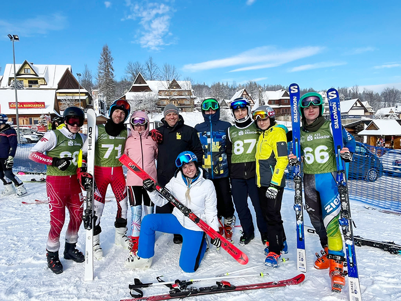 Grupa uśmiechniętych narciarzy pozuje do zdjęcia z trenerem. W rękach trzymają narty. Ubrani są w sportowe stroje narciarskie. W tle zimowy krajobraz. 