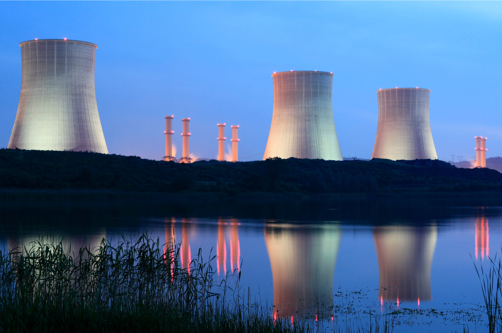 Elektrownia atomowa, widok nocą. Trzy duże kominy i mniejsze. Wszystkie podświetlone. Kominy odbijają się w lustrze wody na pierwszym planie